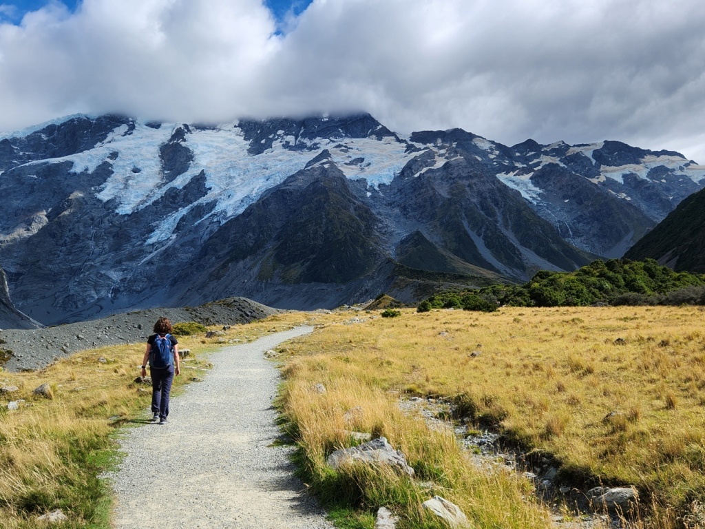 Kaitlyn is walking along a gravel path with her back to camera. Fields of long yellow grass at her side and a large mountain in the distance with a glacier at it's top.